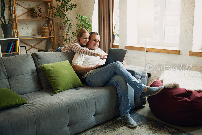 女人站在男人身后，手指着丈夫坐在客厅沙发上拿着的笔记本电脑屏幕。