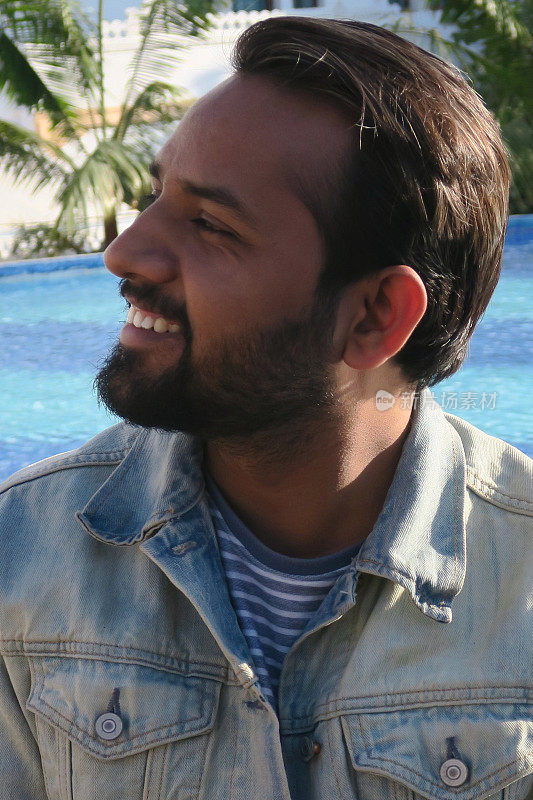 英俊的年轻印度男子坐在游泳池在阳光下看起来快乐的暑假，牙齿微笑，大笑，好看的印度男子享受阳光无限游泳池蓝色马赛克瓷砖，度假模式在牛仔夹克