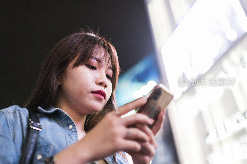 低角度年轻女子使用手机在明亮的夜晚购物区
