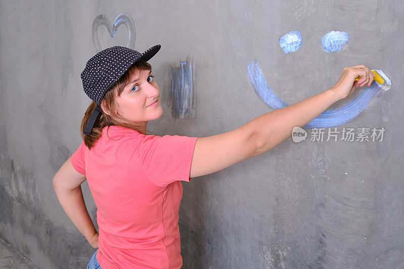 一个戴着帽子的女人在墙上画了一个微笑，特写。油漆工用白漆漆墙。你自己修理新房子。