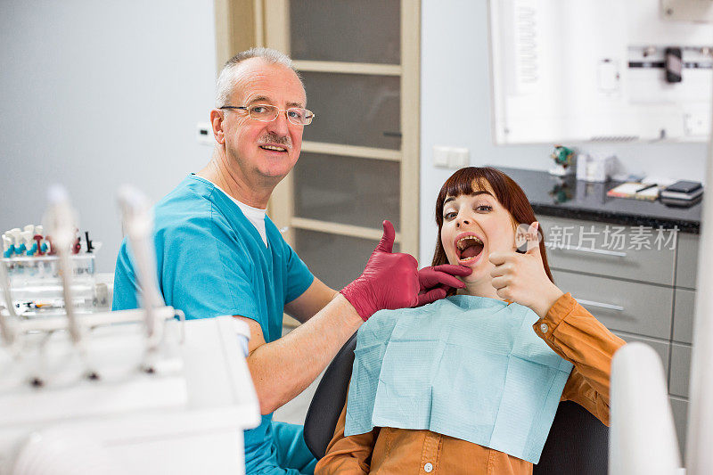 美丽的年轻女子在牙科诊所满意后，成功的牙科治疗，坐在椅子上，显示拇指与资深男医生牙医
