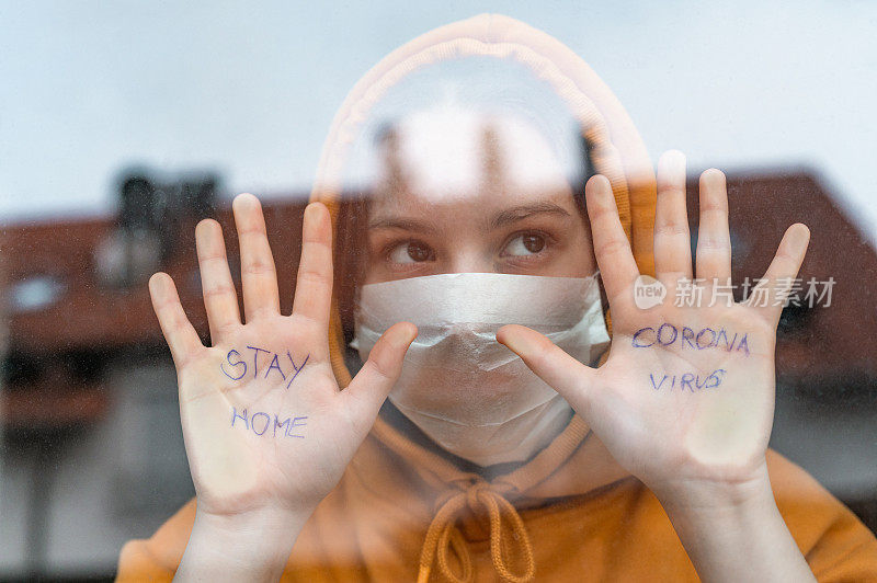 一个十几岁的女孩站在窗前，手上写着“待在家里”和“冠状病毒”