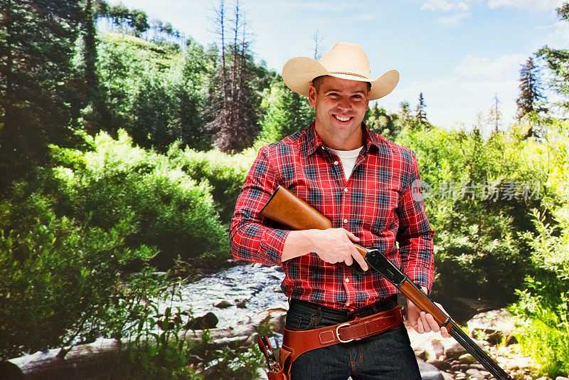 白种年轻男性牛仔在山景前，戴着牛仔帽，手持武器，使用步枪
