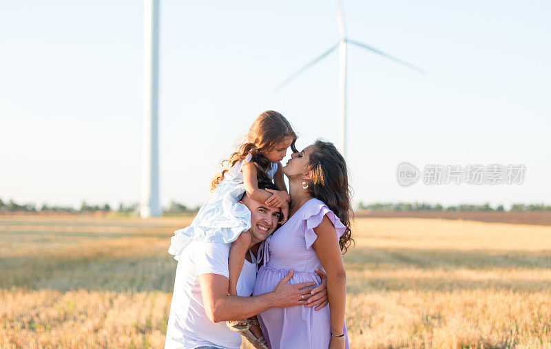 年轻的丈夫，怀孕的妻子亲吻女儿在父亲的肩膀上的田野，风力涡轮机的背景