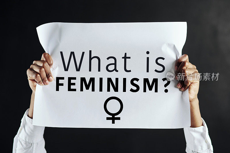 摄影棚里，一个面目全非的女人举着一个牌子，上面写着“什么是女权主义?””
