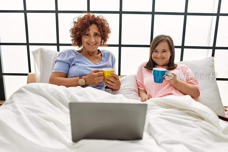 母亲和唐氏综合症的女儿舒舒服服地躺在床上，喝着咖啡，用笔记本电脑看电影