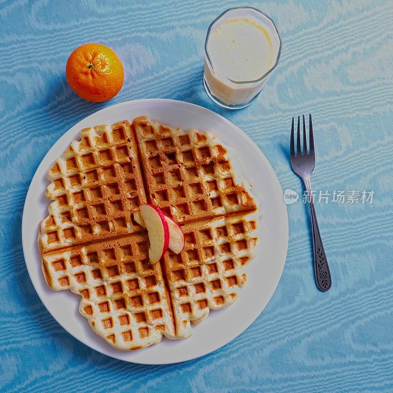 白色盘子上烤苹果华夫饼，上面放着苹果片，配上橘子和一杯蛋酒，蓝色桌布为背景