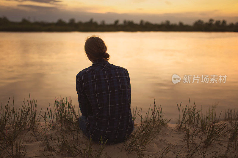 美丽的夏日夕阳背景上的男子剪影。橙色的天空。那家伙正坐在河边。人坐在河岸上