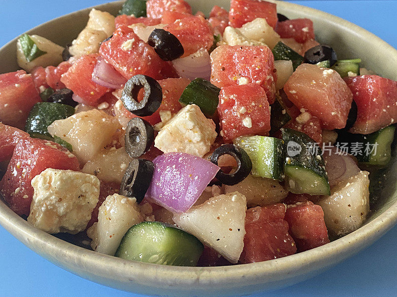西瓜沙拉在碗，切片黄瓜，红洋葱，羊乳酪块，薄荷叶和黑橄榄，蓝色的背景，重点在前景