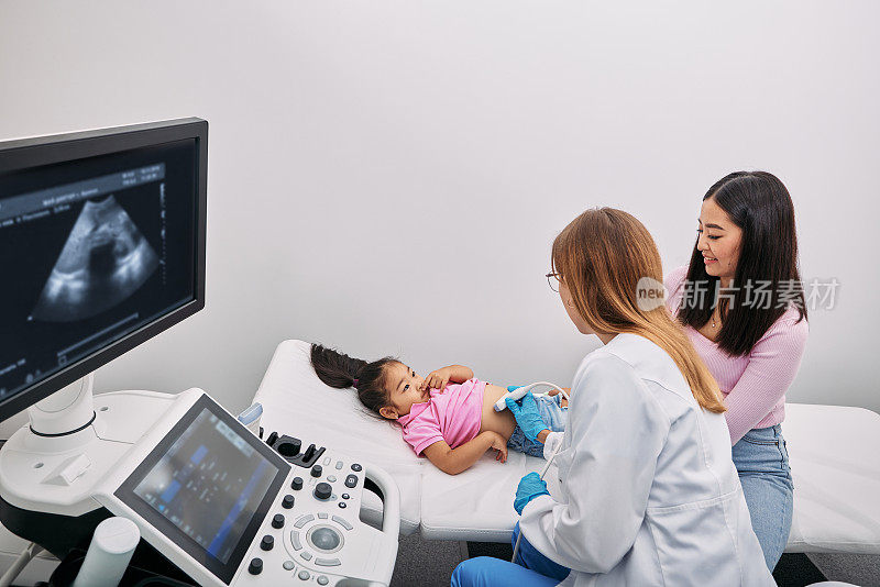 医生用超声探头用超声机扫描亚洲儿童的腹部和内脏器官。小女孩在母亲的支持下，接受腹部超声检查