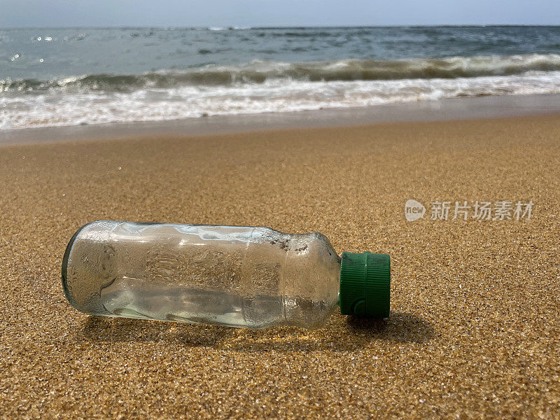 特写图像，空的，透明的塑料饮料瓶与绿色盖子被冲到沙滩上在退潮水的边缘，破碎的海浪冲刷海洋垃圾和岸上的污染，乱扔的沙子，肮脏的海滩，关注前景