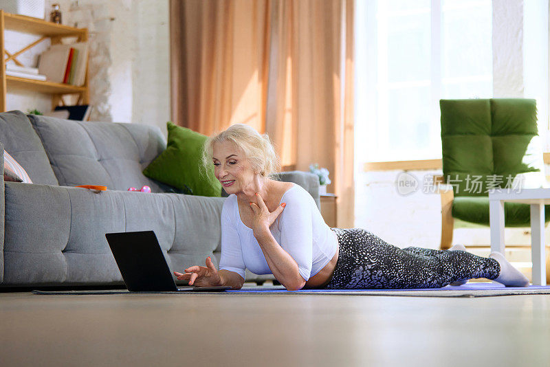 漂亮的高级女性躺在健身matt上，看着笔记本电脑，搜索在线健身视频课程。家教