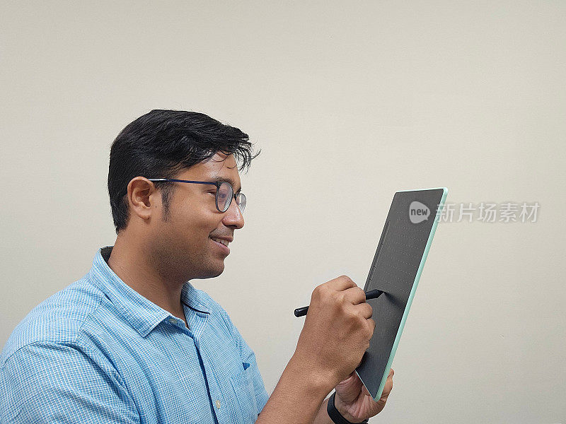 一名印度男子戴着眼镜，穿着蓝色衬衫，在灰色背景下用铅笔拿着平板电脑
