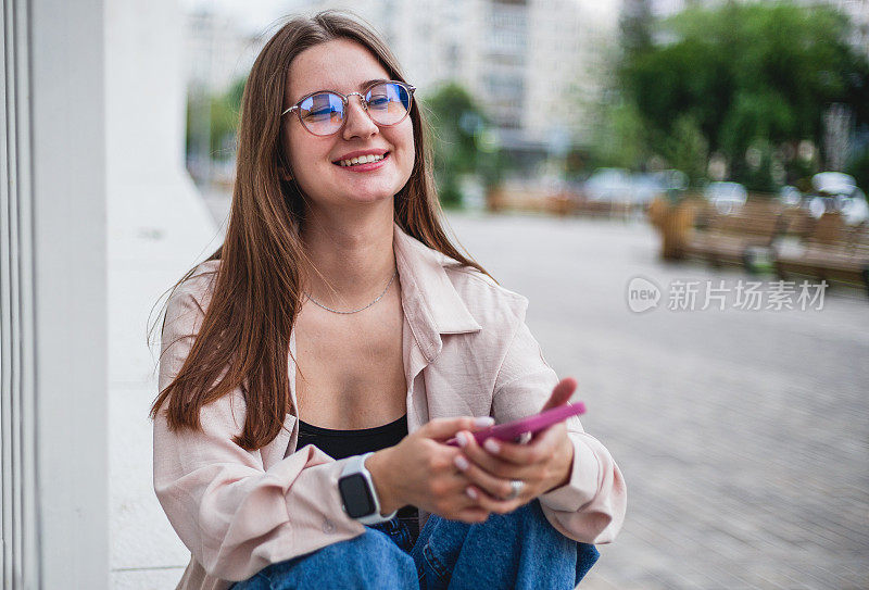 一个穿着牛仔裤、衬衫、拿着手机的年轻女孩坐在街上微笑着
