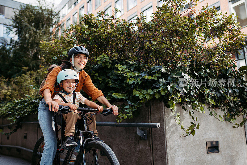 母亲抱着儿子坐在儿童自行车的车架上，座位上，两人都戴着头盔。母亲带着年幼的孩子骑着自行车在城市里穿梭。