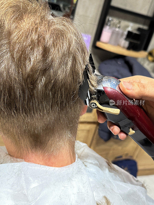 白人男子在理发店理发的特写照片，理发师拿着电动剃须刀，剪头发的人剃短的背部和两侧，理发斗篷，后视图，重点在前景