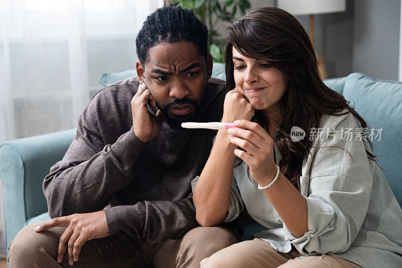 期望，想要或不想要的怀孕。年轻夫妇男女坐在家里的沙发上拿着验孕棒看着。新的生活理念，等待阳性或阴性的怀孕结果