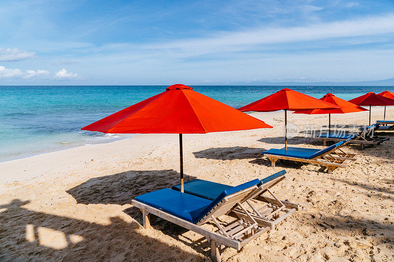 巴厘岛，阳光海滩逃生:充满活力的红色雨伞，金色沙滩上的蓝色躺椅，清澈的海水和蔚蓝的天空。