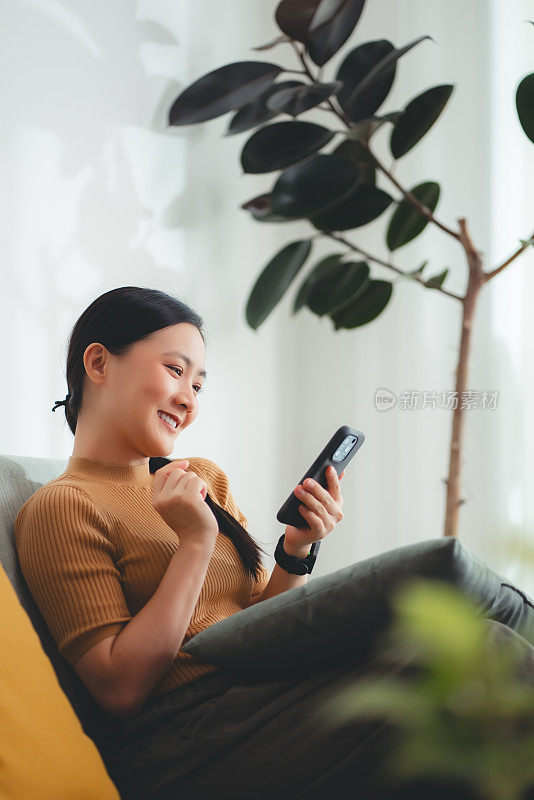 亚洲女人开心地微笑着坐在沙发上，在家里用智能手机。