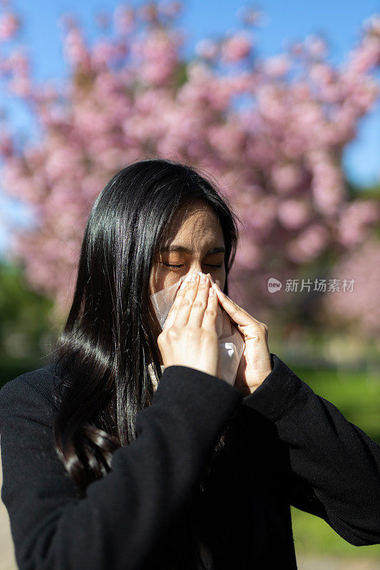 一位年轻的亚洲女子在春天的公园里打喷嚏，在盛开的鲜花中与过敏作斗争。