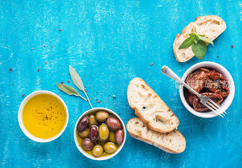 地中海的零食。橄榄、油、晒干的番茄、香草和切成薄片的意大利脆皮面包涂在蓝色的背景上