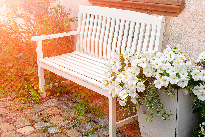 一张舒适的白色木凳，在夕阳的照射下，一个大木盒里长着白色的矮牵牛花。小镇舒适，私人空间