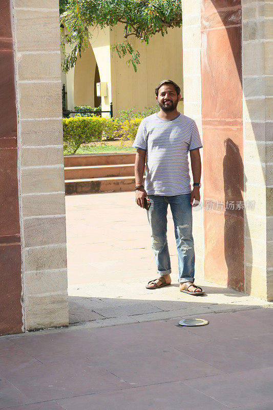 一个20岁出头的年轻印度男子站在柱子旁，看着镜头摆姿势，留着棕色短发，头发梳到脑后，这是一个来自印度德里的帅气男子，穿着蓝白条纹t恤和蓝色破洞牛仔牛仔裤。