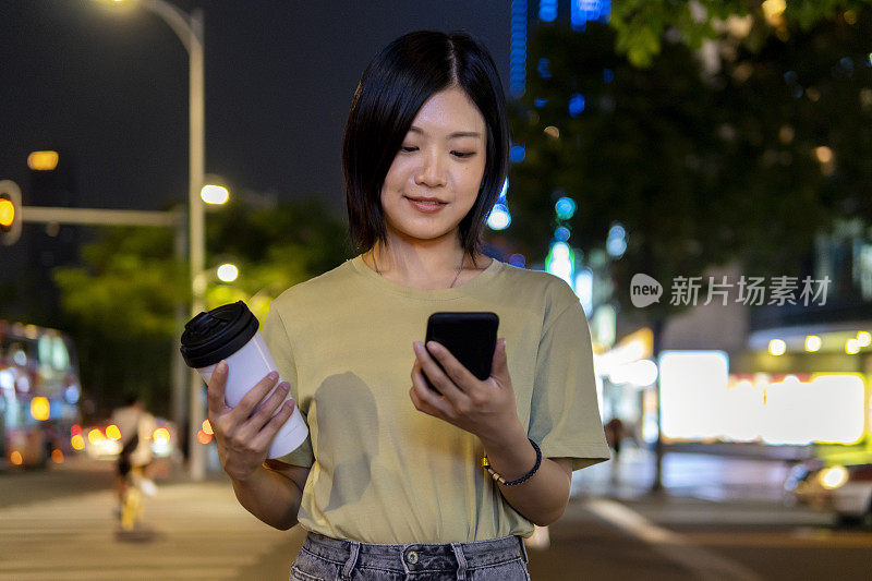 一个美丽的亚洲女人在繁忙的城市街道上用手机端着一杯咖啡