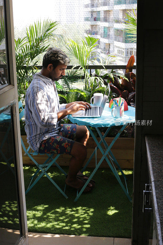 一名男子坐在阳台露台的蓝色圆桌旁，在阳光下用笔记本电脑工作，从走廊厨房门口观看，冠状病毒的新常态概念
