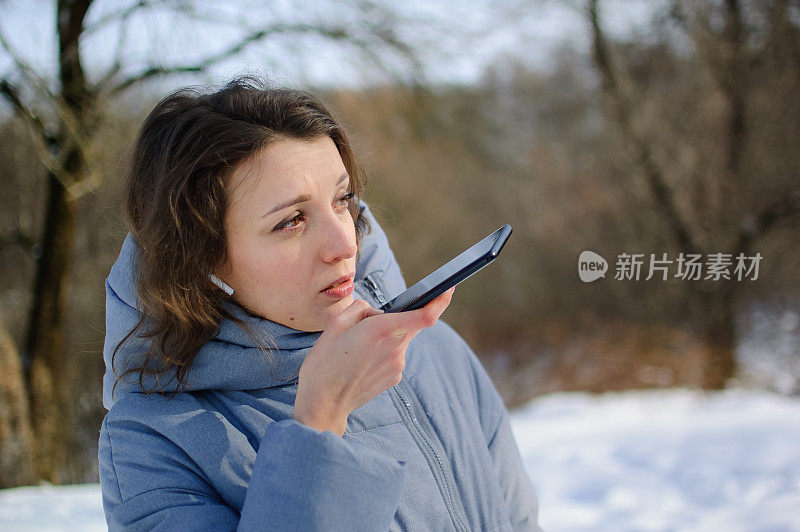 一位穿着蓝色外套的女士正在发送语音信息，解释在室外与移动助手通话的智能手机上的一些事情。用智能手机语音识别和听写的女孩