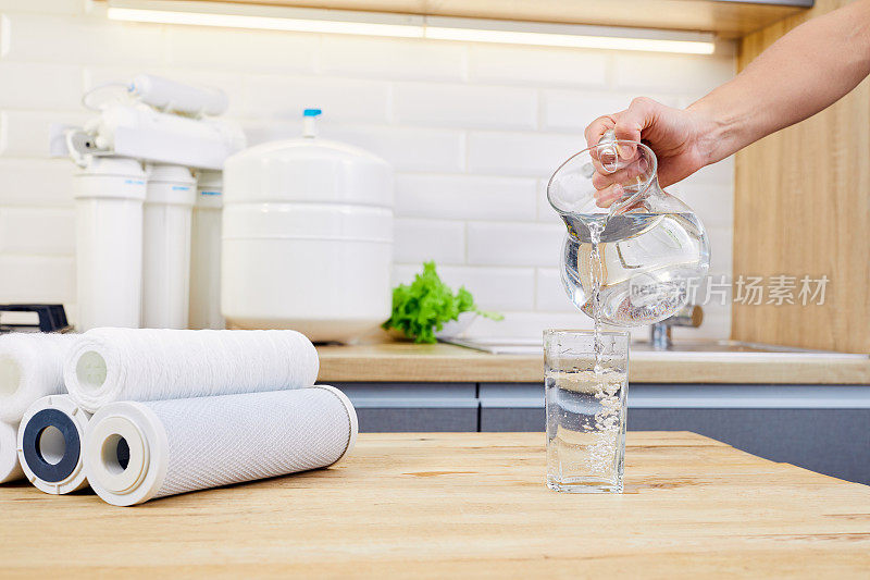 在厨房里，一名妇女正用手将水罐中的纯净水倒入玻璃杯中。背景上的反渗透过滤器。医疗和美丽。Hydratation。