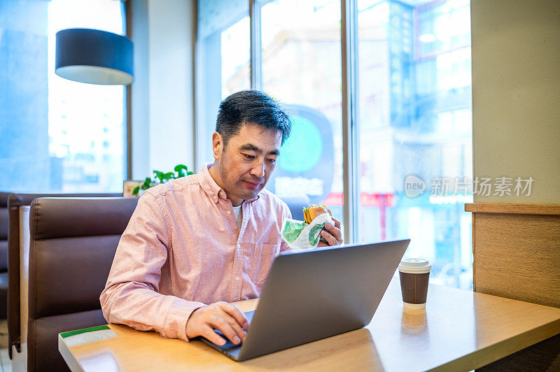 亚洲中年男子在咖啡店吃汉堡用笔记本电脑