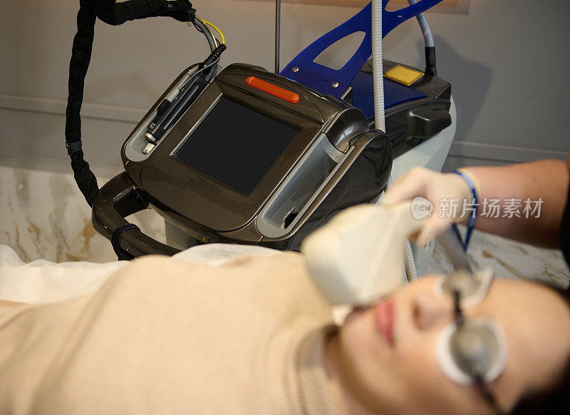 专注于现代激光仪器在医疗水疗诊所模糊白人妇女得到头发去除治疗她的脸在前景