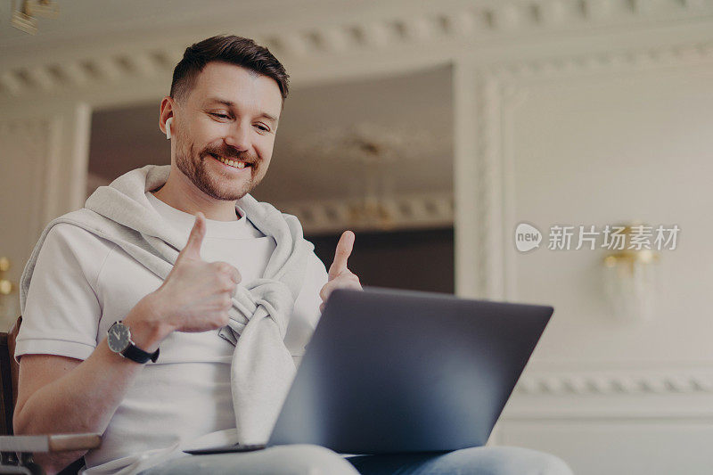 开朗的大胡子男模特有视频会议使okay手势竖起大拇指与朋友在网上交谈穿着便装使用无线耳机笔记本电脑愉快地聊天。