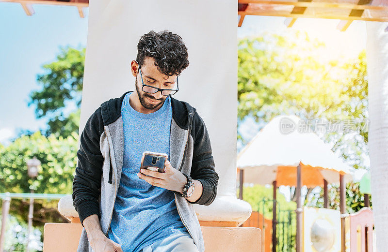 微笑的帅哥靠在公园的墙上用手机，戴眼镜的帅哥靠在墙上用手机聊天，青少年男性靠在墙上发短信