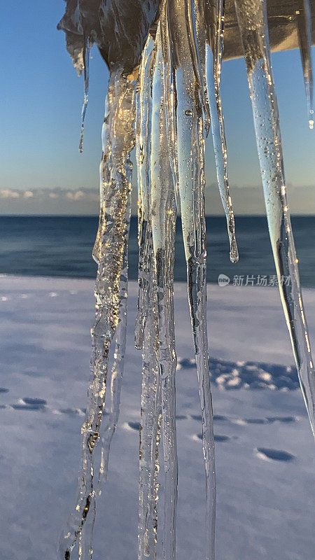 透明的冰柱在特写中悬挂下来，阳光反射在大海上，雪海滩上有脚步声