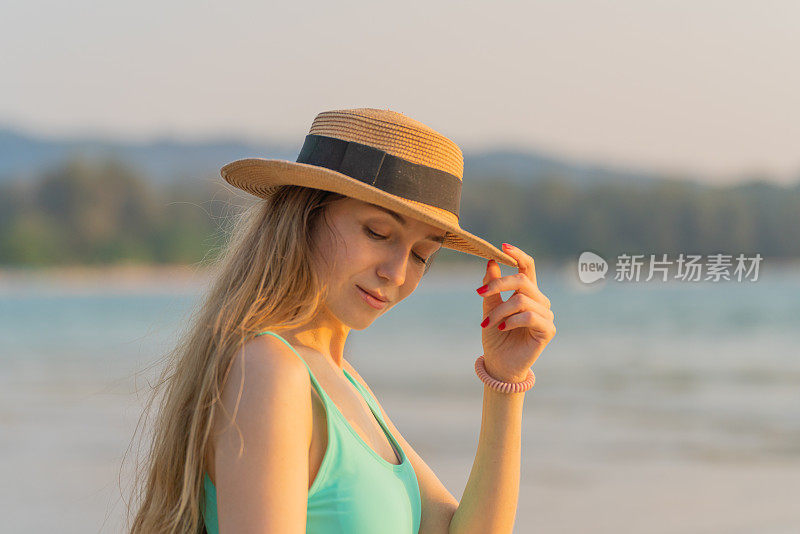 美丽的白人女子在比基尼拿着沙滩帽在海滩上悠闲地摆姿势，同时享受海风和日落的景色