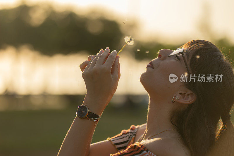 日落时分，年轻的女士正在吹一朵蒲公英花。