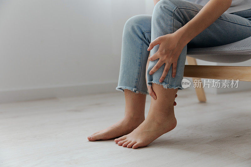 痛苦的腿小腿遭受晒黑美丽的年轻亚洲妇女触摸脚踝在家里室内客厅的特写镜头。伤害，不良健康，疾病概念。酷炫报价横幅