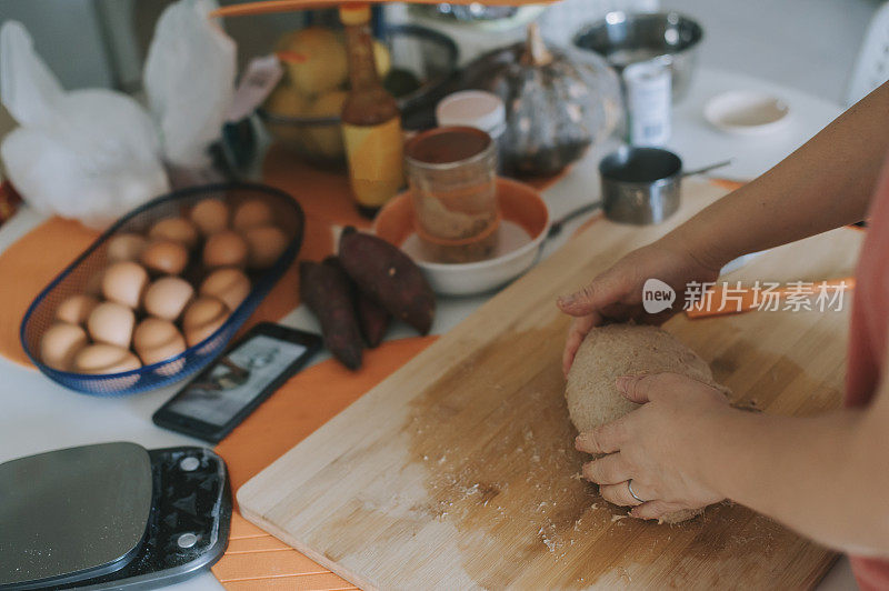 一位亚洲华人家庭妈妈一边准备面团，一边用电话上网查询菜谱和在线课程指导