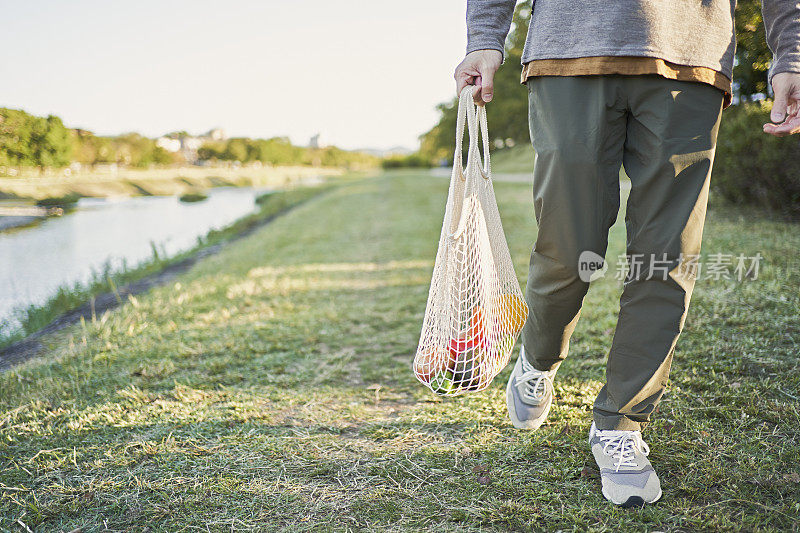 一名日本男子背着装在网眼袋里的杂货，沿着河边的木板路行走