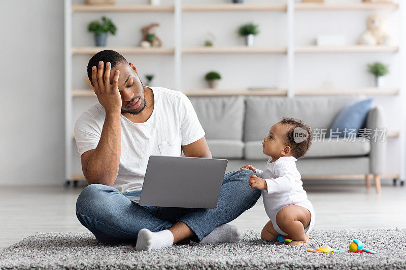 压力大的黑人男子试图用笔记本电脑工作，而婴儿分散了他的注意力