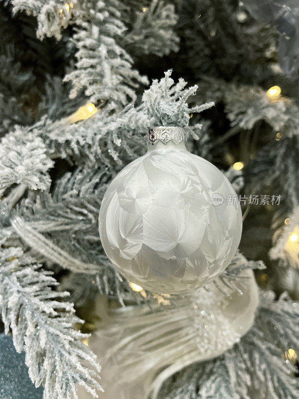 个人白色的图像，霜图案的小饰物挂在人造圣诞树的树枝上，云杉针覆盖着人工喷雾雪，模糊的白色精灵灯的背景