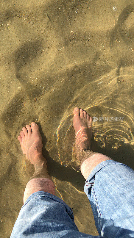 一个白色的英国男人的脚在海里划水，卷起的蓝色牛仔牛仔裤在金色的沙滩上浸湿浅浅的海水，海浪拍打着海岸的海岸线，光着脚尖，涟漪，倒影，夏日海滨男人的腿站在海里