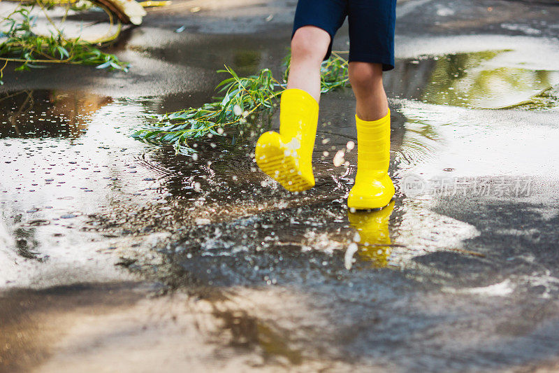 快乐的孩子。夏雨。水坑在沥青。穿着黄色雨靴的孩子。孩子跑过水坑。寒冷的秋天的雨。潮湿多雨的天气。水飞溅。倒影在水中