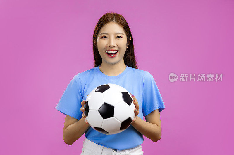 快乐年轻的亚洲女子孤立粉红色背景看着相机，拿着一个足球，国际足联世界杯概念