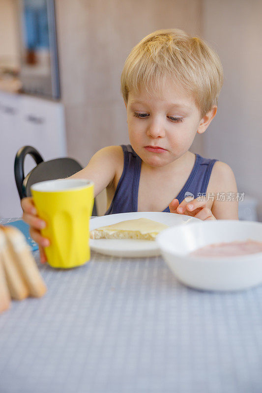 金发小孩在餐桌上吃自制早餐，拿着水杯