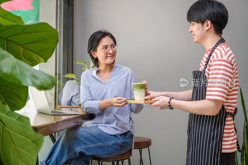 年轻的亚洲男子服务员在咖啡店的桌子上为他的顾客提供咖啡