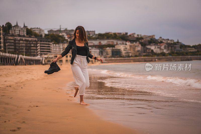 年轻时尚的女性在日落时赤脚走在海滩上抱着她的海岸