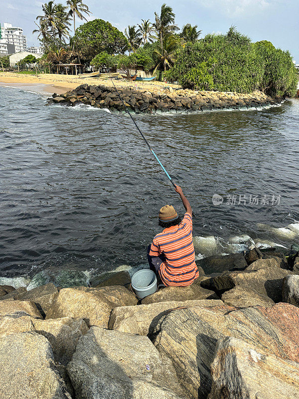 渔夫坐在海岸线上的岩石自然海防上的鱼竿特写图像，海岸侵蚀抛石岩石装甲墙，海岸管理，岩石堆，海滩岩石装甲，海浪，背景中的城市摩天大楼，科伦坡，斯里兰卡